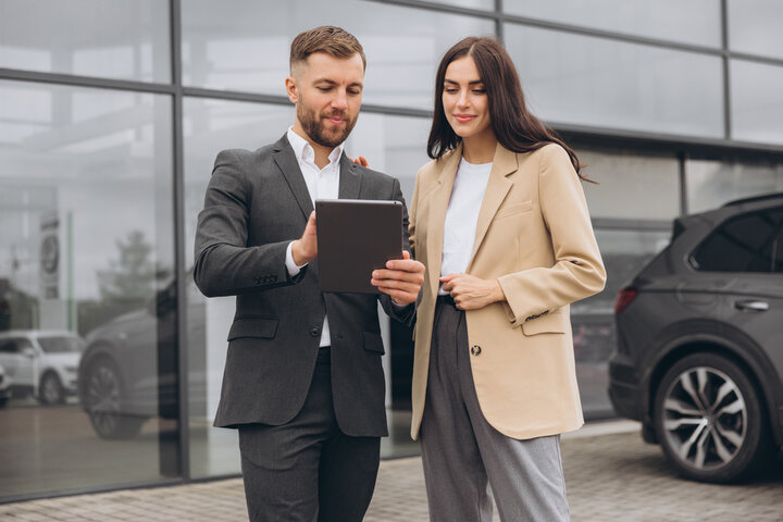 Bearded business man car salesman in grey suit giving explanations on tablet to pretty young woman in beige jacket outside car dealership