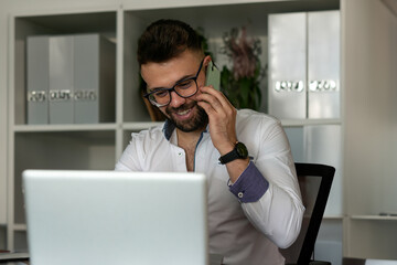 Smiling male employee working on laptop talking on cell